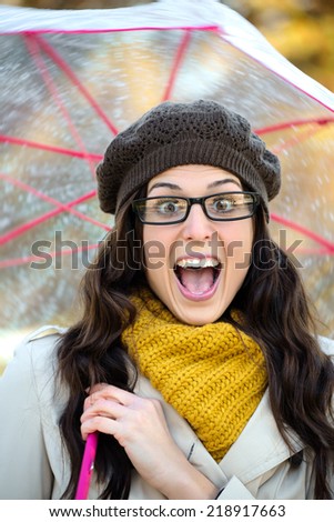 Surprised happy woman in autumn having fun playing with umbrella under the rain. Funny brunette with mouth open wearing glasses and scarf.