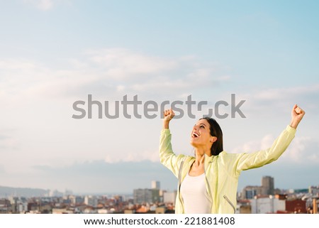 Happy woman celebrating fitness and sport exercising success. Successful female athlete raising arms to the sky on city skyline background.