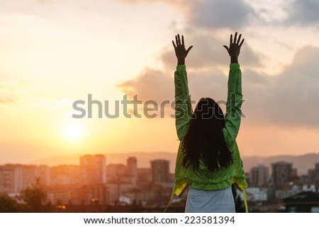 Back view of a happy woman celebrating sport goals and fitness lifestyle success. Female athlete raising arms to the sky after exercising towards beautiful sunset or morning over city skyline.