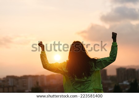 Back view of a successful woman celebrating sport goals and fitness lifestyle success. Female athlete raising arms to the sky after exercising towards beautiful sunset or morning over city skyline.