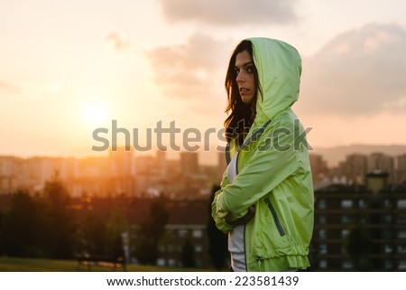 Serious female athlete crossing arms towards sunrise or sunset over city skyline for sport motivation and fitness lifestyle concept.