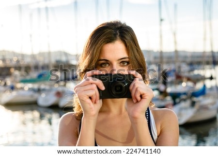 Young woman on summer vacation travel taking photo by the harbor in Gijon, Asturias, Spain. Tourist pointing camera.