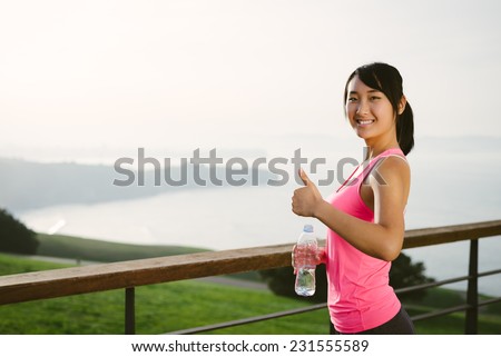 Positive happy female athlete taking a workout break for drinking water. Asian woman on a training rest towards the sea. Motivation and healthy lifestyle concept.