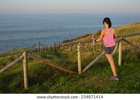 Back view of female asian athlete doing leg stretching exercise after running by the sea. Sporty woman warming up on beautiful nature background. Fitness and healthy lifestyle concept.