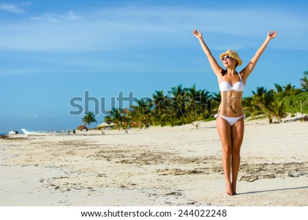Beautiful blissful woman in white bikini enjoying tropical beach and caribbean summer vacation. Tanned brunette raising arms and enjoying freedom by the sea at Playa Paraiso, Riviera Maya, Mexico.