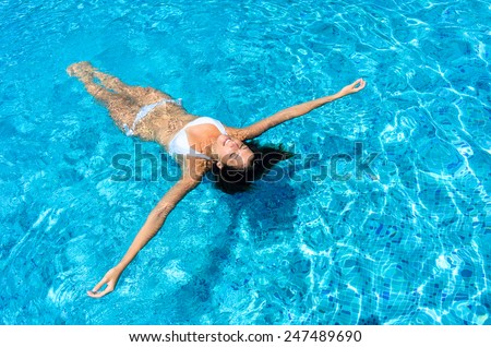 Young woman relaxing in swimming pool on summer vacation. Brunette caucasian model in white bikini floating in water.