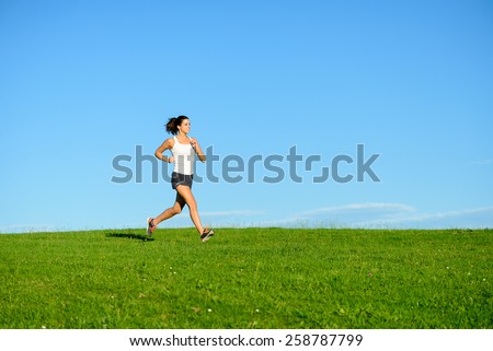 Woman running on summer or spring grass field. Female athlete exercising outdoor. Brunette fit sporty model training towards clear blue sky background.