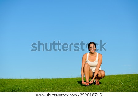 Sporty woman ready for running and exercising workout outdoor at grass field. Female brunette athlete lacing footwear for training towards clear blue sky copys pace background.