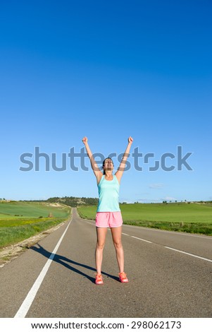 Happy woman celebrating running and sport success. Female athlete raising arms to the sky during training on countryside road.