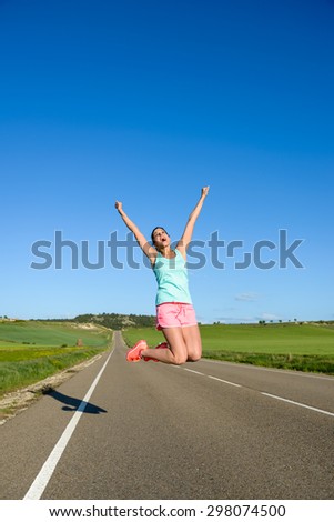 Happy woman jumping for celebrating running and sport success. Female athlete raising arms to the sky during training on countryside road.