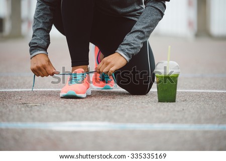 Fitness workout and healthy nutrition concept.  Detox smoothie drink and running footwear close up. Female athlete tying sport shoes laces before training outdoor.