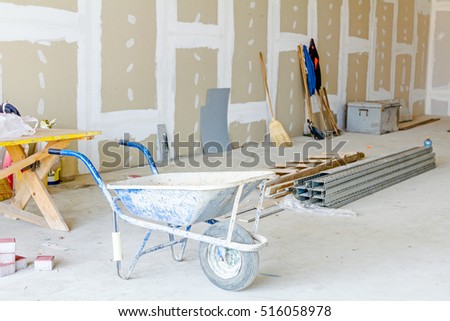 Wheelbarrow is in front of tools for ground work. Thin square metal profiles for dry wall are stacked on ground in background.