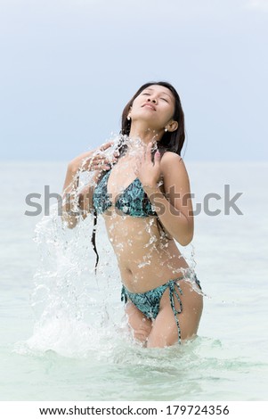 Young woman in a bikini splashing in the sea spraying droplets of water over her body as she stands with her head tilted back in enjoyment