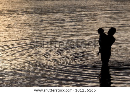Father hugging his son in the sea silhouetted at sunset in an intimate embrace enjoying the tranquility of the evening with ripples spreading over the surface of the water around them