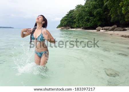 Young woman in a bikini splashing in the sea spraying droplets of water over her body as she stands with her head tilted back in enjoyment