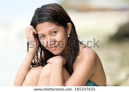 Beautiful charismatic young Filipina woman sitting with her knees bent up to her chin on a beach looking at the camera with a gentle  smile