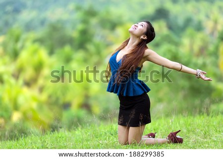 Beautiful stylish Filipina woman rejoicing in nature kneeling in the green grass in the countryside with her head back and arms outspread in the sunshine with a joyful smile