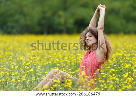 Beautiful young woman stretching her arms high in the air with a smile of pleasure and satisfaction as she sits in a rural meadow full of colorful yellow flowers, with copyspace