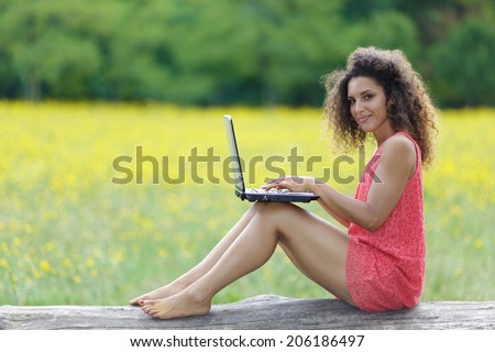 Pretty barefoot woman with frizzy curly hair wearing a trendy miniskirt sitting on a fallen tree trunk in a rural field working on a laptop and looking at the camera with a friendly smile