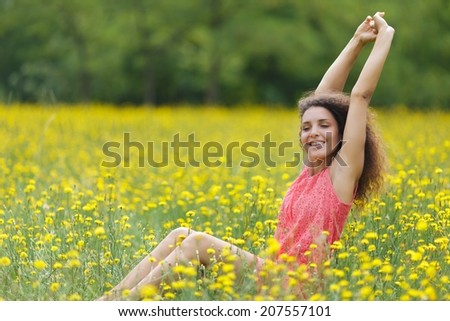 Beautiful young woman stretching her arms high in the air with a smile of pleasure and satisfaction as she sits in a rural meadow full of colorful yellow flowers, with copyspace