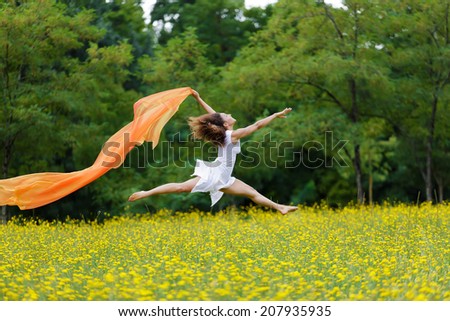 Agile barefoot woman with curly brown hair leaping in the air in a meadow of yellow wildflowers trailing a colorful orange scarf in the breeze as she celebrates her freedom and the beauty of nature