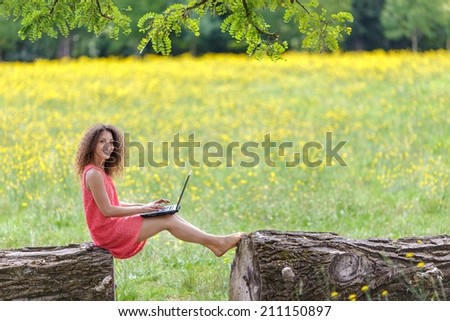 Young woman using laptop in nature sitting on a log in a dandelion summer field