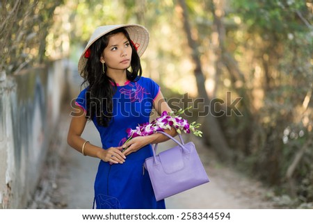pretty young Vietnamese woman in a stylish blue outfit carrying flowers as she walks through a tree-lined avenue in the park