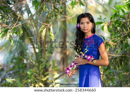 Gorgeous young Vietnamese woman standing holding sprays of fresh tropical flowers as she stands in a park with leafy green trees