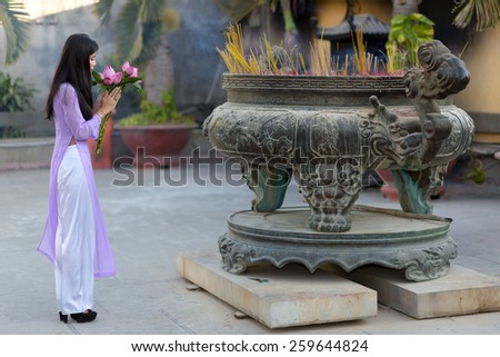 Young Asian woman making a votive offering of fresh pink flowers at an open-air Buddhist shrine filled with burning sticks or incense