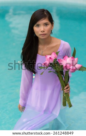 Pretty Vietnamese woman in a flowing lilac dress standing in water holding a bouquet of fresh flowers and looking at the camera