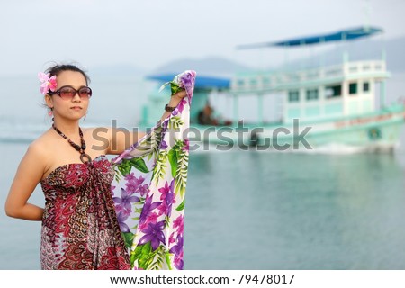 asian woman holding sarong posing at sea with boat at the background, thailand