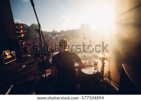 Open air view from the stage to the drummer and another band mates in sunset warm lights. Stage smoke, sun rays in it. Silhouettes of the summer festival