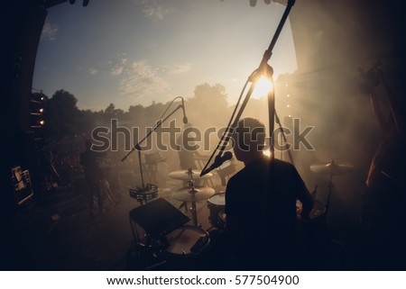 Open air view from the stage to the drummer and another band mates in sunset warm lights. Stage smoke, sun rays in it. Silhouettes of the summer festival