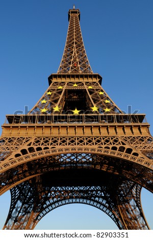 Eiffel Tower lit by golden sunlight against clear blue sky, displaying the European Union Circle of Stars symbol on the front.
