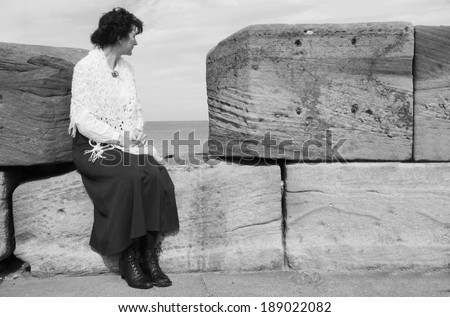 Black and white tinted photograph of a woman in Edwardian dress sitting on Whitby Pier, looking out to sea.