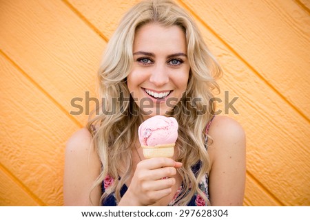 A cute twenty something woman has fun while eating her strawberry ice cream cone while standing in front of an wooden bright orange wall.