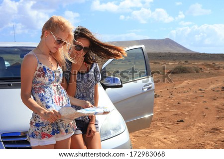 Two young women with car look at road map with mountain landscape in background Two young women with car look at road map with mountain landscape in background