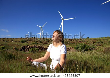 young blond woman doing yoga on the grass on a wind farm beneath eolic generator