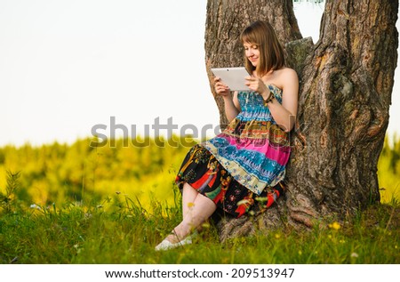 young beautiful woman sitting in a field under a tree and uses a tablet, for study, for watching movies, chtneiya books, web browsing