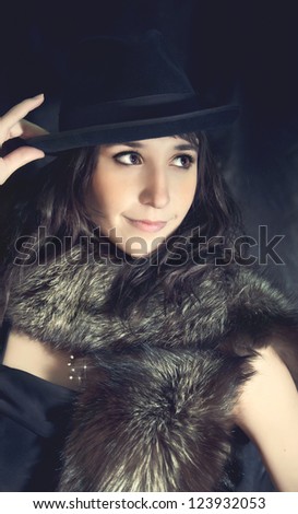 glamorous brunette girl with a fur collar and a black hat posing on black background