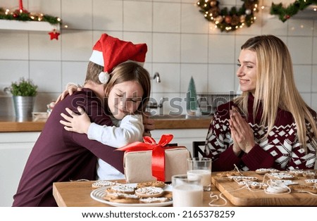 Dad in santa hat presenting Christmas gift to kid daughter hugging at home. Happy father making present greeting cute child girl with xmas gift sitting at kitchen table celebrating family holiday.