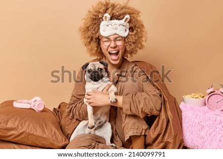 Emotional woman with curly hair exclaims loudly dressed in slumber suit poses with pug dog poses on bed in bedroom wrapped in soft blanket surrounded by alarmclock bowl of cornflakes headphones