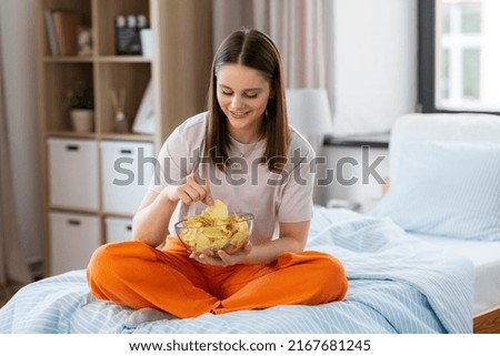 fast food and people concept - happy girl eating crisps sitting on bed at home