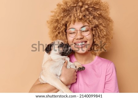 Happy curly haired woman receives kiss from favorite pet smiles broadly have fun together wears transparent glasses and t shirt stand indoor. Pug dog licks owner with love. Display of affection