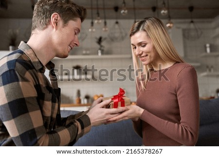 Happy young woman receiving gift box from loving man at home. Young couple in love celebrating Valentines day enjoying romantic date holding present box surprise sit on couch together.
