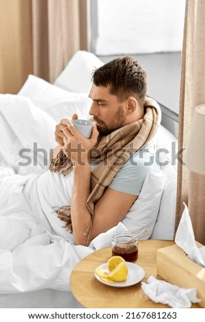 health, illness and people concept - sick man drinking hot tea with lemon and honey in bed at home