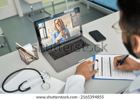 Over shoulder view of videocall chat with female older patient and indian male doctor in modern clinic hospital using laptop computer, consulting online remotely. Telemedicine healthcare concept.