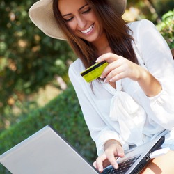 Young elegant woman wearing straw hat and white dress holding credit card with laptop sitting on bench at park and looking away. She is shopping or making payments carefree online