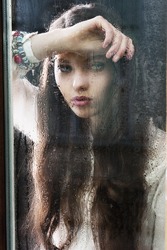 Portrait of a lovely young lady looking through glass window - Indoor in a dark cloudy day, she looks down at left, her head is resting on the right arm and the left hand is near the chest