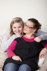 Mother sitting in a couch being hugged by happy teenage daughter. Mother is looking to her daughter, daughter is looking happily into the camera.